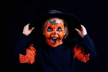 Siyah şapkalı, balkabağı makyajlı bir çocuk ellerini şapkaya uzatır ve siyah bir arka planda yüksek sesle güler.,