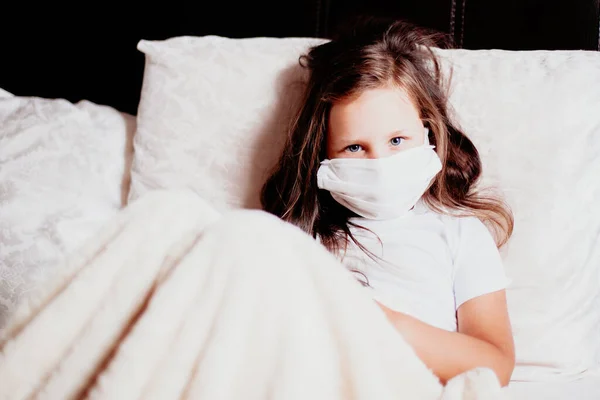 Девушка сидит в белой маске на кровати в спальне, самоизоляция от простуды, вторая волна вирусов — стоковое фото