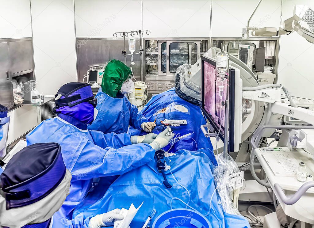 This shot shows an ongoing emergency coronary angioplasty procedure perform in a cardiac catheterization laboratory during COVID. The medical staff is in full PPE.