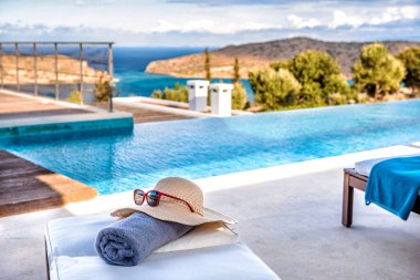 Tahta güneş yatağı terasta duruyor, güverte sandalyesinde şapka ve mavi havlu, mavi gökyüzü ve arka planda beyaz bulutlar