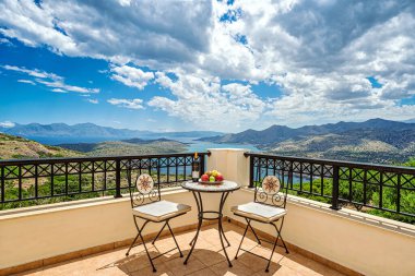 Masa ve balkon / terasın köşesinde iki sandalye. Girit adasına muhteşem bir panoramik manzara, Akdeniz denizi ve beyaz bulutlu mavi gökyüzü. Masanın üzerinde meyveler ve bir şişe şarap var..