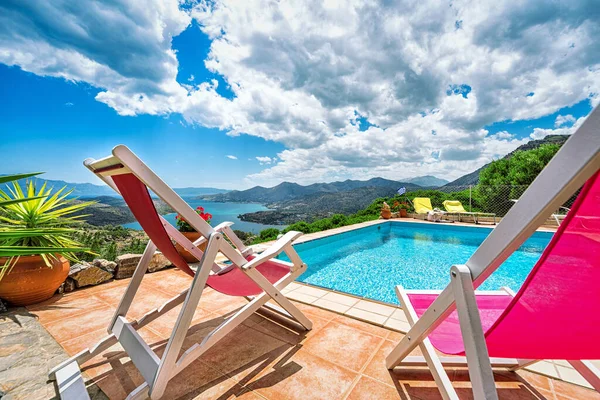 Pool terrace with two sun chairs in pink and white overlooking the amazing panoramic view to the island with its blue sky and white clouds, blue watter of the pool and amazing sea and mountains