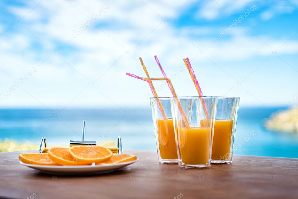 Tasty fresh orange fruit juice in glasses with straws and the plate with fresh orange slices on the brown surface. The background is blurred blue sky and sea.