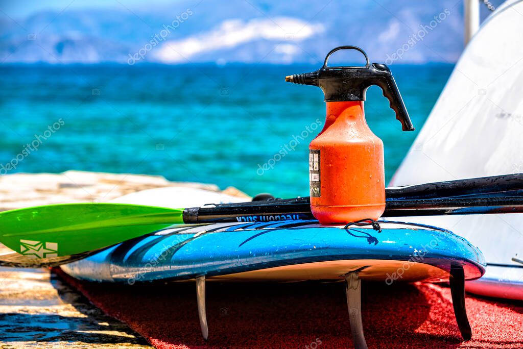 Concept of disinfection of water sport equipment. Atomiser bottle standing on the sup board and paddle near, sea and sky at the background.