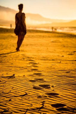 Dalgalı ve basamaklı kumsal, arka planda bulanık kadın silueti inanılmaz günbatımının tadını çıkarıyor. Altın saat fotoğrafı.
