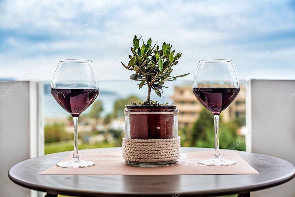 Small olive tree in the pot on the middle of the table and two glasses with red wine at the sides, blurred backgroud with sky and clouds, balcony or terrace concept.