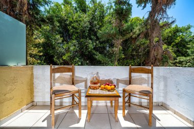 Balkona bir bak. İki ahşap sandalye ve sepetinde sürahi ve meyve olan küçük bir masa var. Arka plandaki yeşil bahçede..