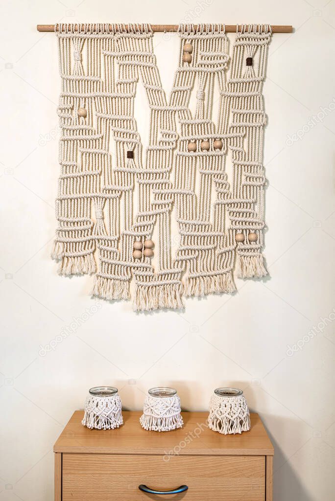 Hand crafted macrame wall decoration with wooden elements. hanging on a wall and below placed three hand made candle holders, all the same size.
