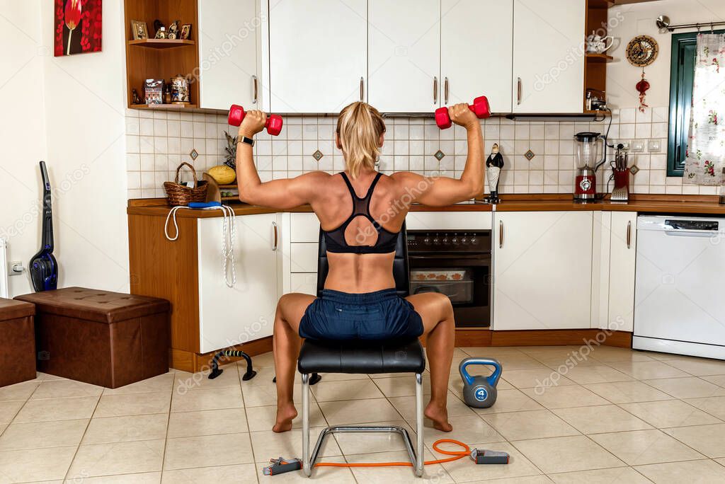 Sporty woman on the chair doing exercise for shoulders with dumbbells, elbows and hands in position up. Kitchen and home workout fitness.