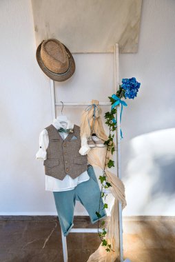 Baby boy suit consisting of green grey pants, white shirt, beige vest, shoes and hat hanging on the decorative white ladder. clipart
