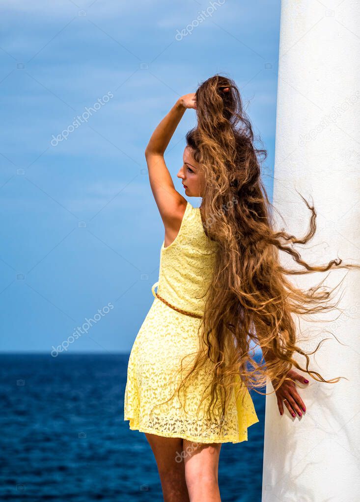 Sexy long hair model in yellow dress, standing with the back, one hand fixing the wild hair