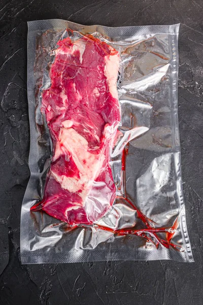 Vacuum packed organic beef chuck roll steak for sous vide cooking on black textured background, top view