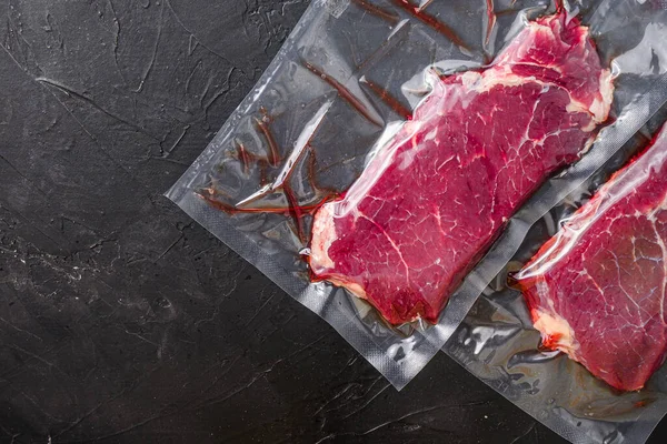 Rump steak in vacuum sealed bag on black textured background, side view space for price.
