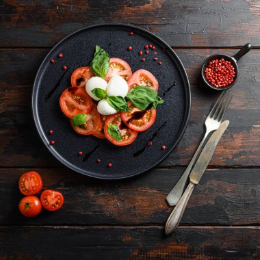 Caprese salatası domates ve fesleğen yapraklı mozzarella dilimleri zeytin ağacının üzerinde koyu masa üstü siyah tabak kare ekin manzarası.