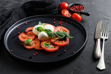 Caprese salatası İtalyan mutfağı konsepti domates ve fesleğen yapraklı mozzarella dilimleri siyah seramik platwantimakarna siyah desenli çatal ve bıçakla yakın plan.