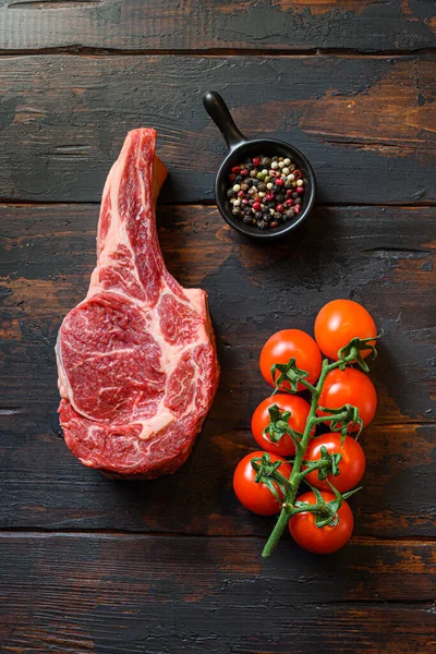 Raw uncooked black angus beef cowboy steak closeup with tomatoes and peppercorns on wooden dark old rustic table planks