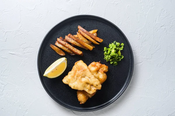 British Traditional Fish and chips with mashed peas, potato fries and slice of leman on round black plate over white concrete textured table.