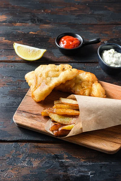 British Traditional Fish and chips with mashed peas, tartar sauce inn crumpled paper cone  on wood chopping board dip and lemon - fried cod, french fries, lemon slices, tartar sauce, ketchup tomatoe served in the Pub or Restaurant over old wooden pla