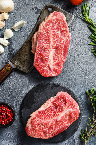 Raw top blade flat Ironcut, on black slate , and meat butcher cleaver marbled beef  with herbs tomatoes peppercorns over grey stone surface background top view close up.
