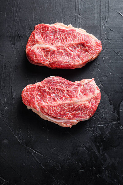 Raw top blade flat Ironcut, on black slate , bio marbled beef  with herbs tomatoes peppercorns overblack stone surface background  top view close up.