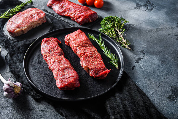 Raw denver, top blade,  tri tip steak on a black plate and stone slate with seasonings, herbs  grey concrete background. Side view close up selective focus new wide angle space for text.