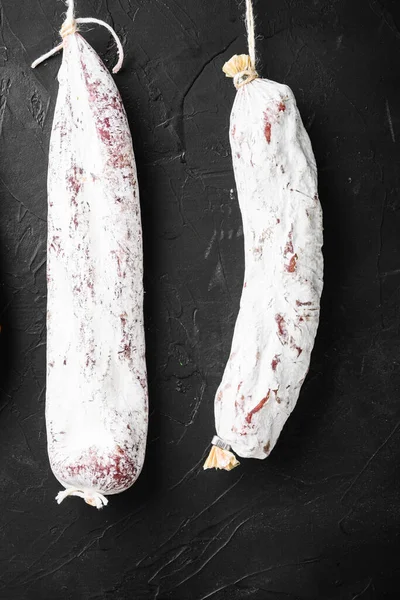 İspanyol salamı, fuet ve sucuk sosisleri siyah arka planda asılı duruyor..