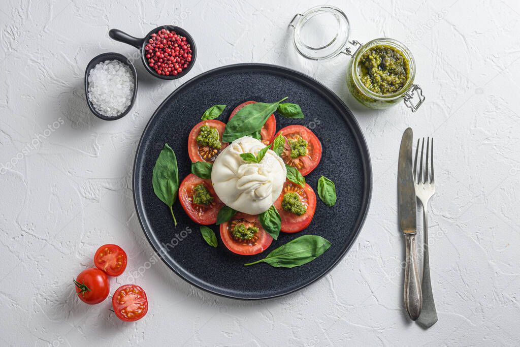 Salad with traditional italian burrata cheese made from cream and milk of buffalo or cow with pesto and tomatoes and seasonings on black flat plate top view flatlay white concrete background  and old fork and knife.