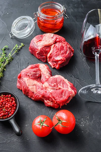 Organic chuck eye roll steak cuts, with red wine glass , rosemary, spicy chili oil on black textured background  side view.