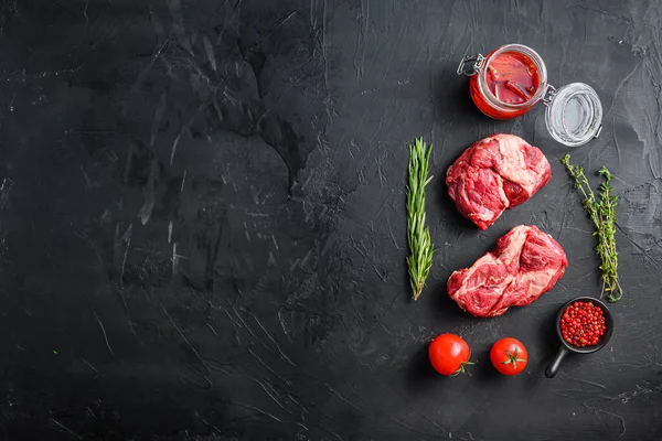 Raw chuck eye roll steak cuts, with rosemary, spicy chili oil and peppercorns  on black textured background  top view with space for text