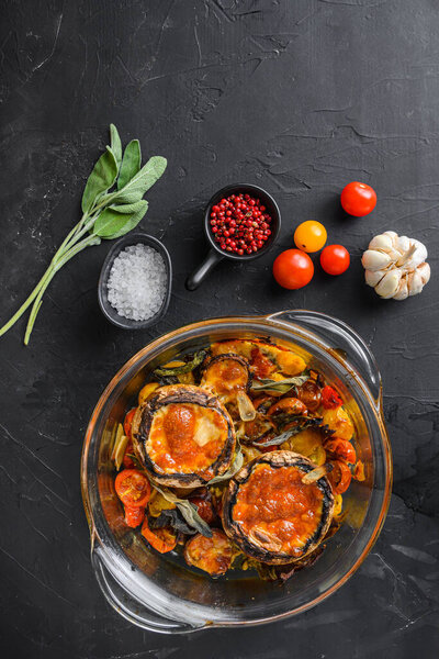 Portobello mushrooms,baked with ingredients  cheddar cheese, cherry tomatoes and sage  in glass pot on  black stone background top view space for text