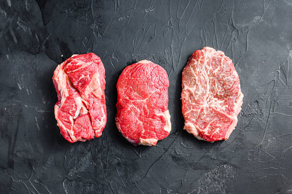 Set of raw  alternative cuts beef steaks, top blade, rump and chuck roll, on black textured background, top view