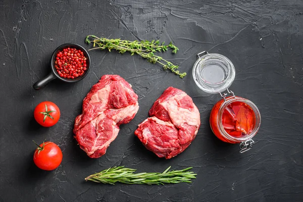 Raw chuck eye roll steak cuts, with rosemary, spicy chili oil and peppercorns  on black textured background  top view