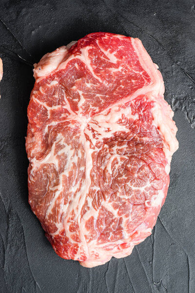 Raw top blade beef steak cut, on black textured background, top view