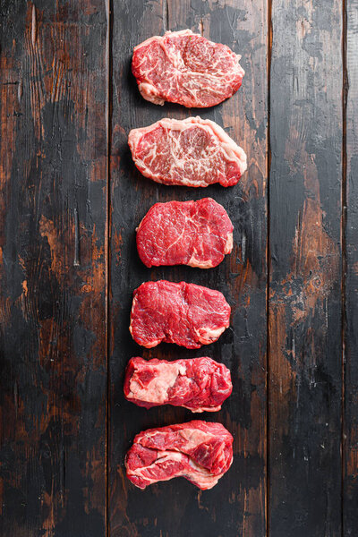 Set of different raw beef steaks on old wooden dark planks top view, space for text