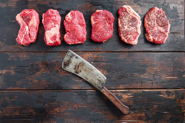 Different raw beef steaks with seasonings and american butcher cleaver on old wooden dark planks top view.