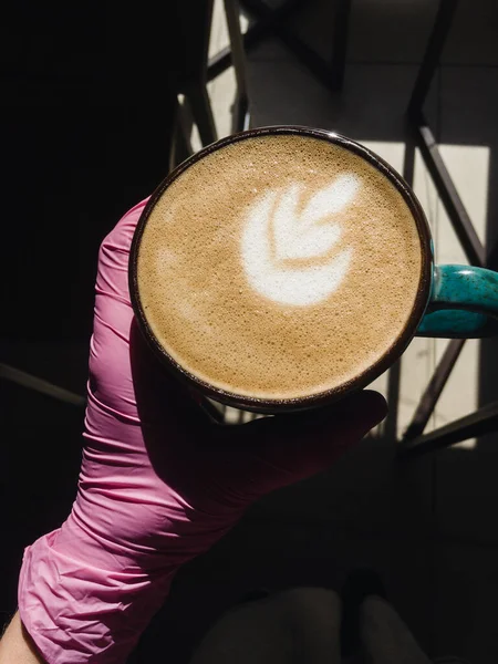 A hand in a pink glove holding a cool green handmade clue cup of hot cappuccino lavender decorated with latte art on a wooden table in a sunny weather