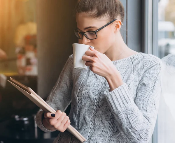 Chica joven mirando a la tableta y sonriendo en la cafetería con ventana grande . — Foto de Stock