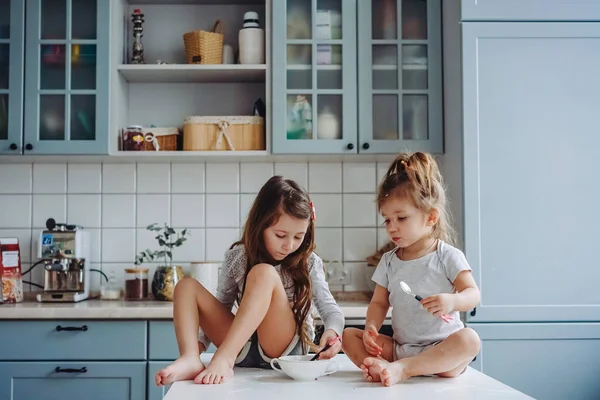 Two little girls in the kitchen sitting on the table. — Stock Photo, Image