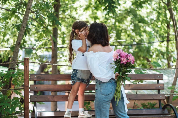 Junge Mutter und Tochter auf Bank mit einem Strauß rosa Pfingstrosen — Stockfoto