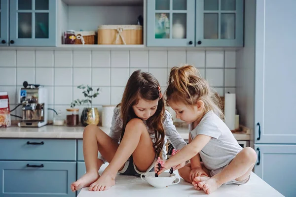 Two little girls in the kitchen sitting on the table. — Stock Photo, Image