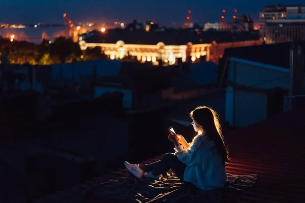 Ung flicka sitter på natten på taket av ett hus. Vacker kväll — Stockfoto