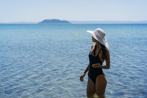 Young girl in a bathing suit and a striped hat is standing in the blue sea — Stock Photo, Image