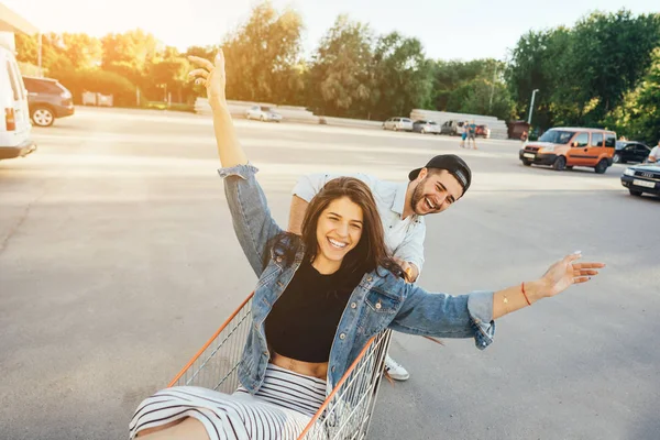 Handsome guy carries his girlfriend in cart in the parking lot — Stock Photo, Image