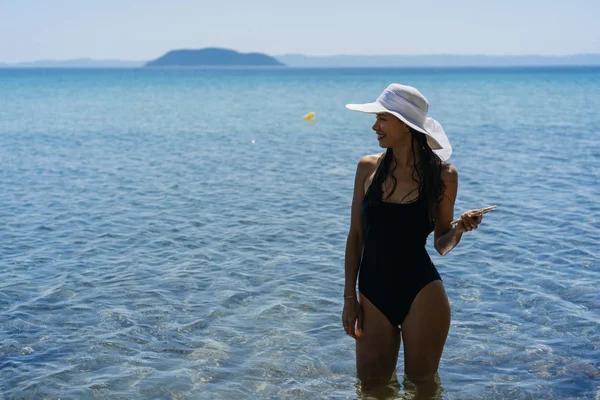 Chica joven en un traje de baño y un sombrero a rayas está de pie en el mar azul —  Fotos de Stock