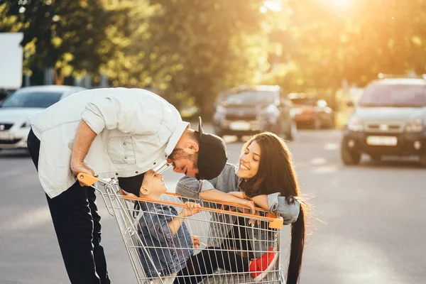 Les jeunes parents mettent leur fils dans un chariot et s'amusent dans le parking . — Photo