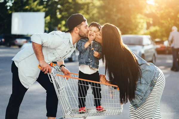 Young parents put their son in a cart and kiss him in the parking. — Stock Photo, Image