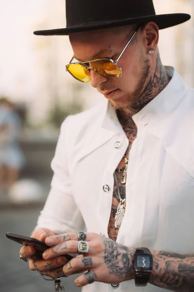 Un joven con sombrero y gafas de sol sostiene un teléfono móvil en sus manos —  Fotos de Stock