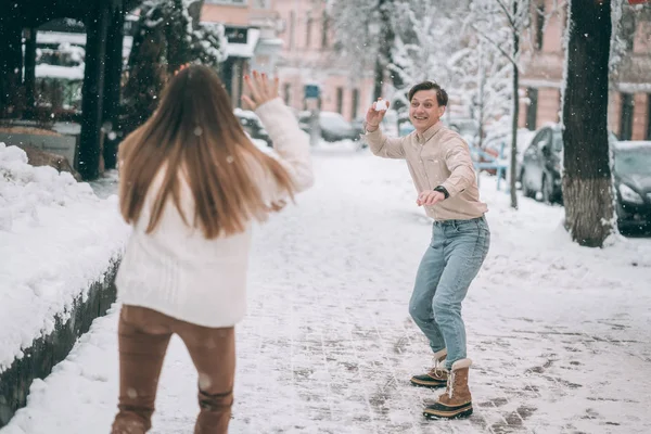 Joyful jovem casal em camisolas está jogando bolas de neve na rua . — Fotografia de Stock