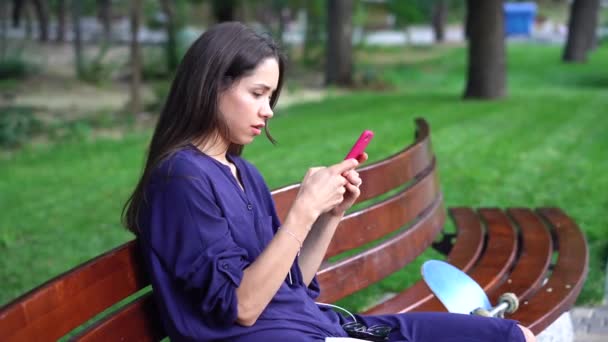 Žena sedí na lavičce a pomocí smartphone přes pozadí park. — Stock video