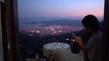 Kadın bir akşam şehrin bir arka plan üzerinde ev cam kenarında oturan bir akıllı telefon kullanır.